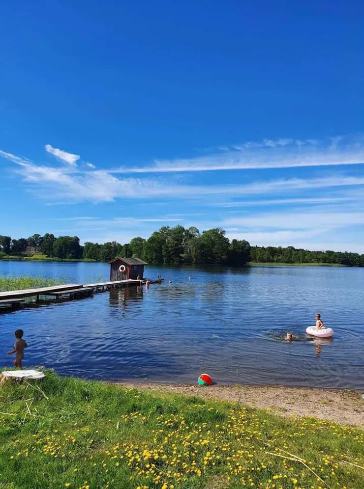 Utsikt över en sjö med klart vatten under en somrig himmel med moln. I förgrunden syns en grönskande strandbank.