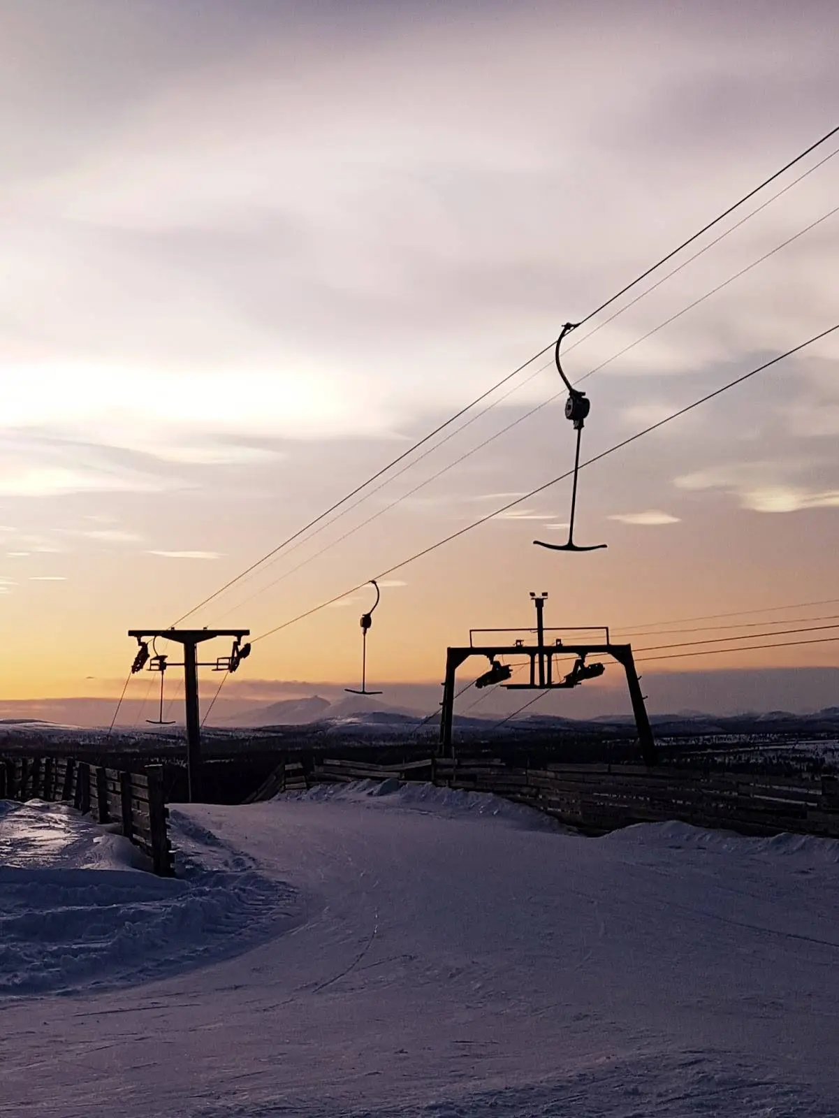 Vy över Idre Fjäll Camping i vintertid, vid skymning. Snö täcker marken och himlen är färgad i varma toner av soluppgång eller solnedgång. Det är kallt och frostigt, med ett stilla och lugnt ljus över platsen.