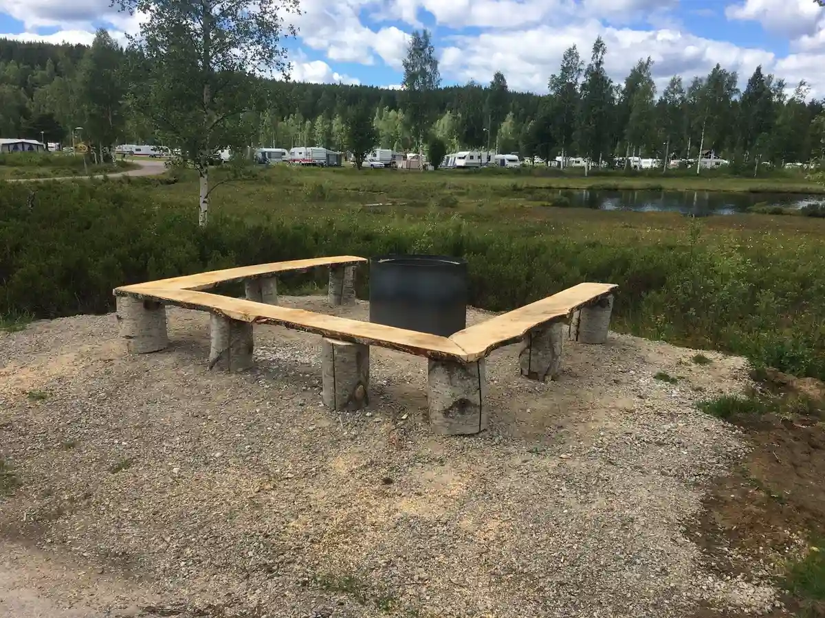 En picknickbänk och bord i trä står på en gräsyta i en campingmiljö, omgiven av grönskande träd.