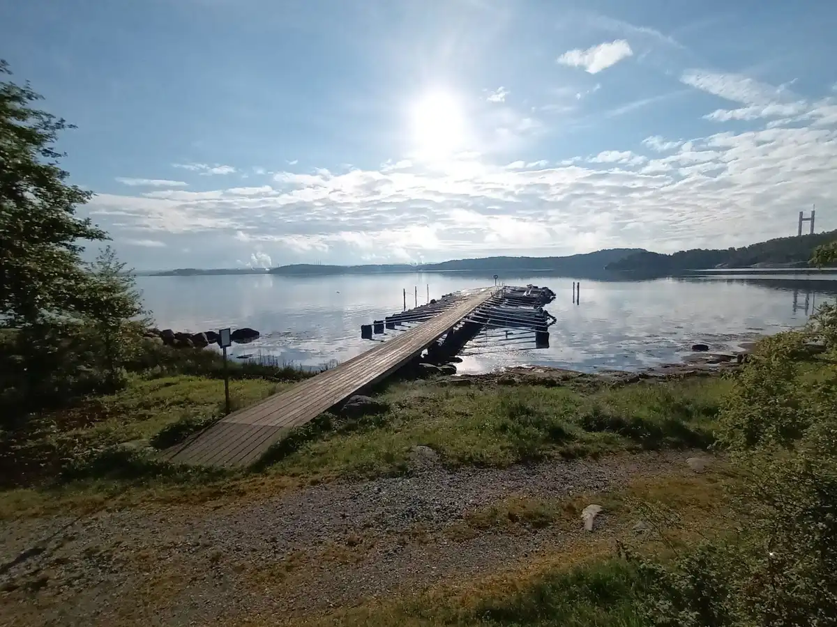 En vacker vy över Almöns bad & camping med en brygga som sträcker sig ut i det lugna vattnet. I bakgrunden syns en sjö omgiven av grönska och ett par moln på himlen.