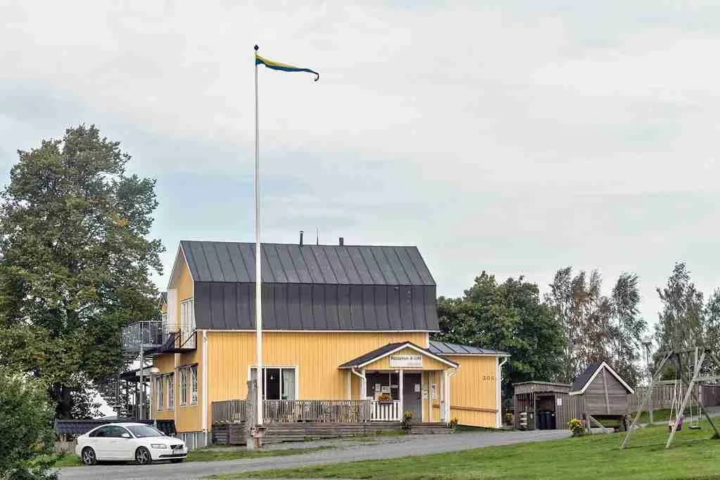 En mysig sommarstuga med röd fasad och vita detaljer, omgiven av grönska. Ett skuggigt grönområde ligger i förgrunden och en gruslagd uppfart leder fram till stugans veranda.