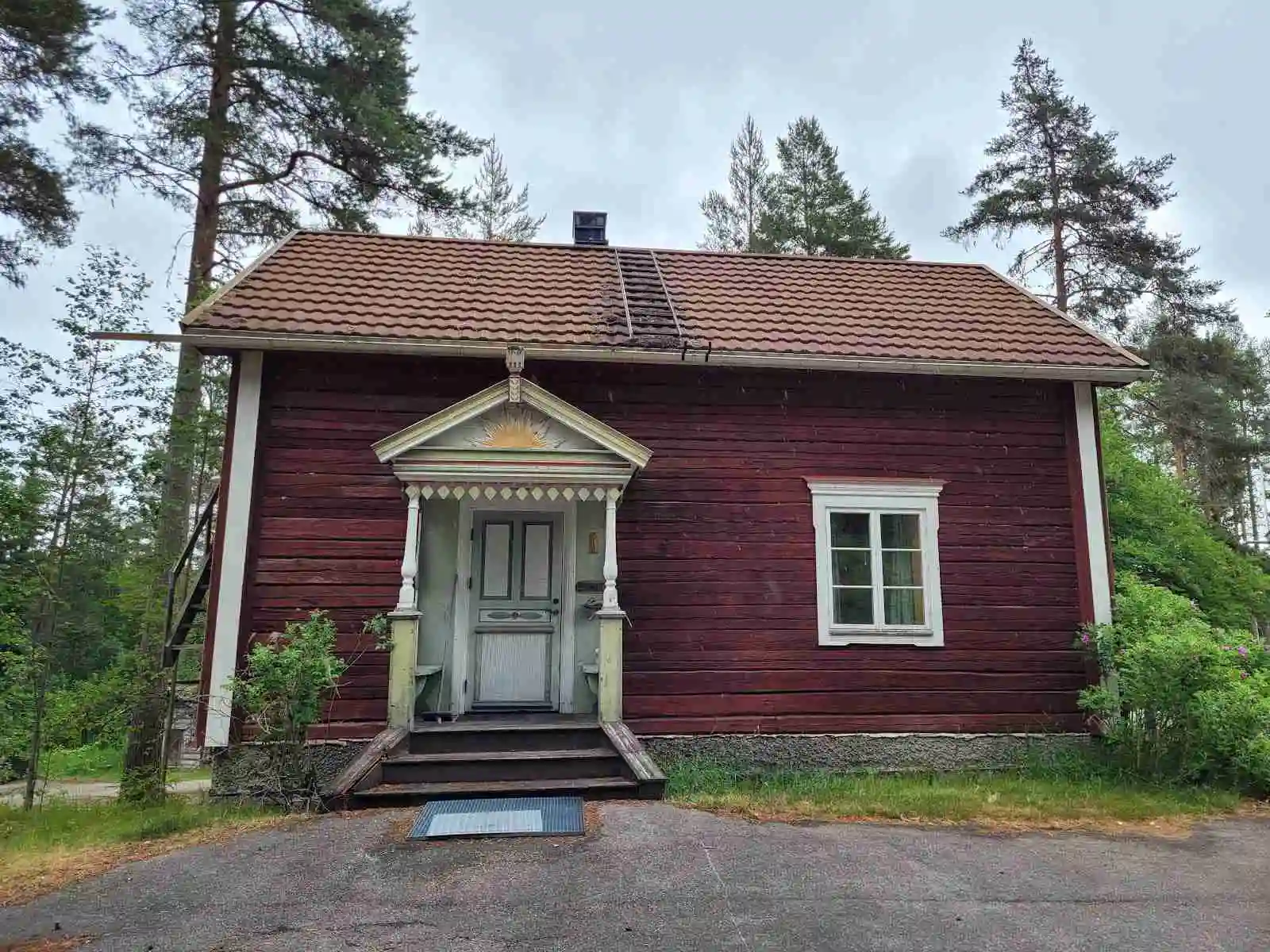 Bilden visar en timmerstuga med ett träskiffertak och en tydligt framträdande träfasad. En trädörr finns vid ingången till stugan, som är omgiven av en idyllisk omgivning vid Skålsjögården Timber Lodge.