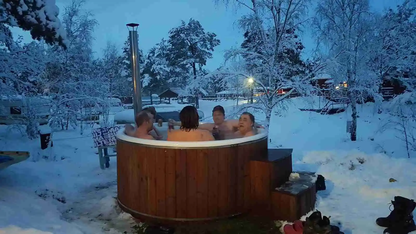 En vinterkväll på Arkösunds camping med snö täckande marken. En uppvärmd badtunna står klar bredvid en stuga, vilket ger en inbjudande kontrast till det kalla vädret. 