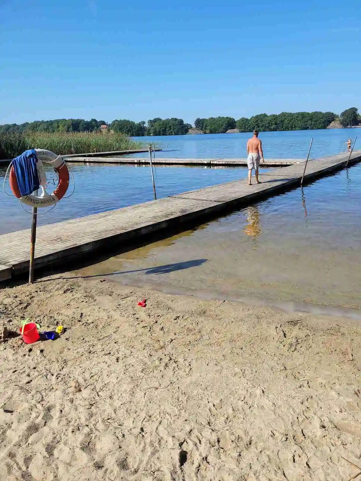 En solig sommardag vid Ivö camping med människor som njuter av sin ledighet. En brygga sträcker sig ut i en lugn sjö, perfekt för rekreation och semesterfirande.