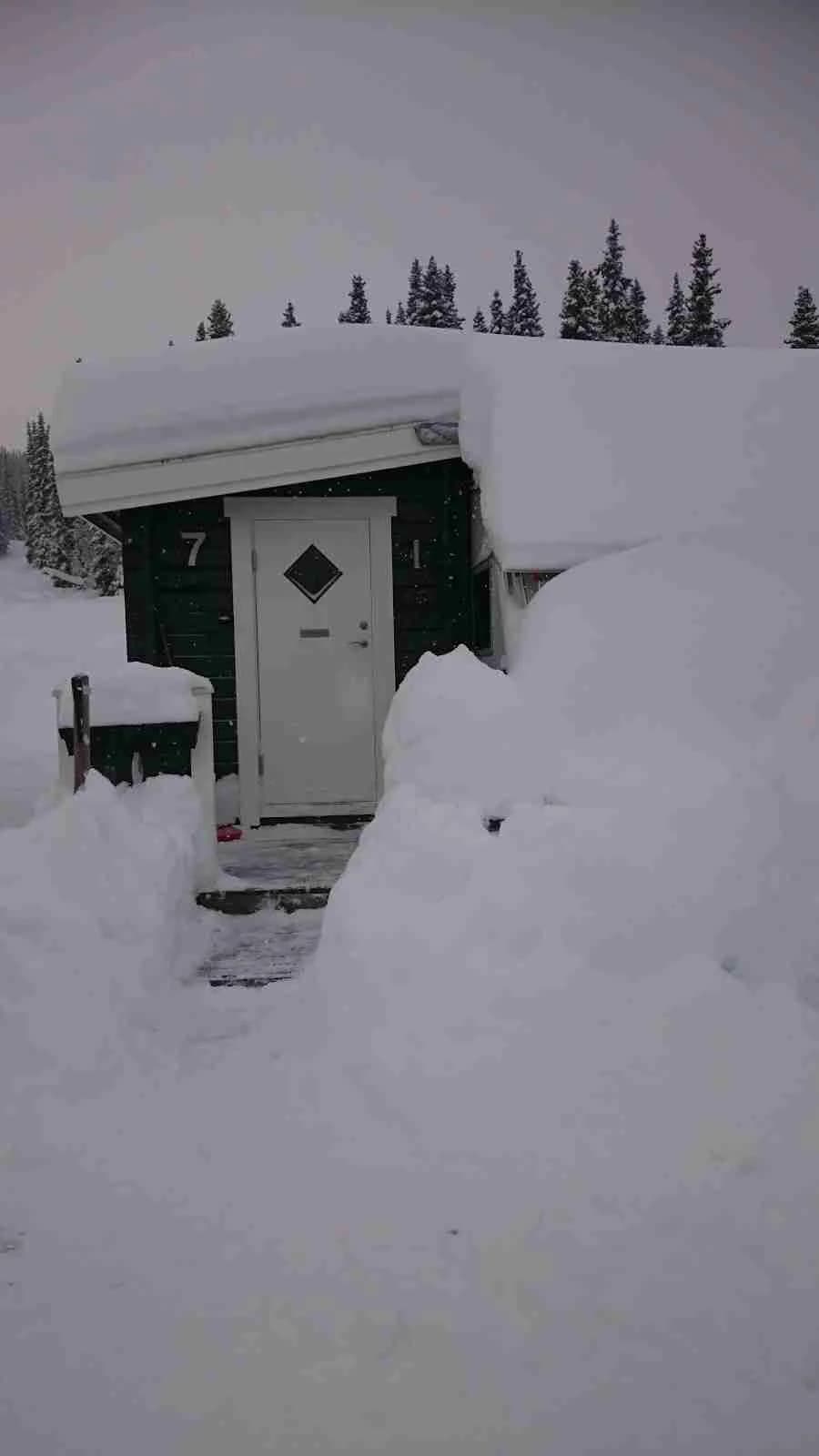 En snötäckt stuga omgiven av barrträd i en vinteridyll, med skidbackar i närheten och ett täcke av is och frost i landskapet vid Sportcamping Åkersjön.