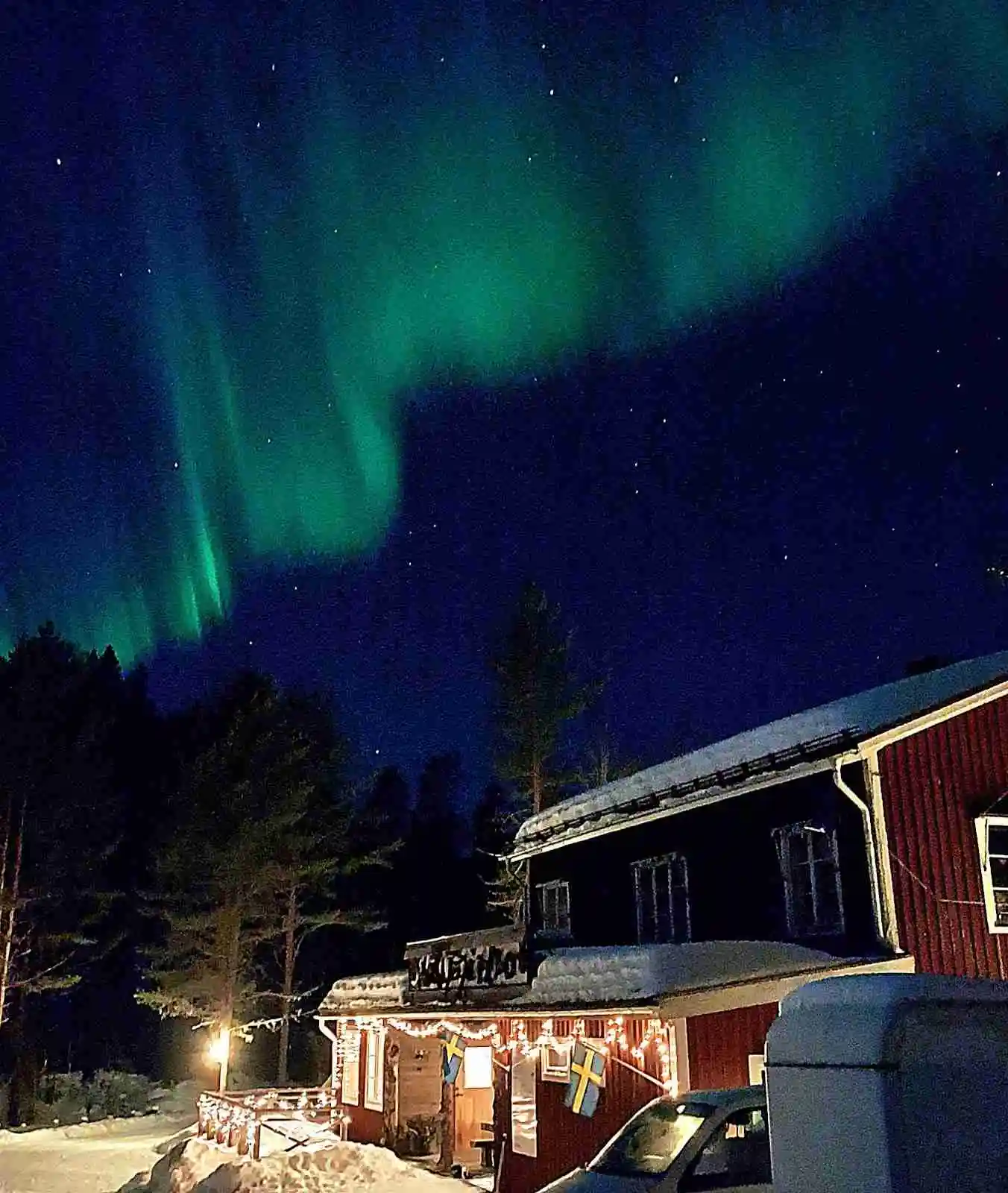 En nattscen vid Skålsjögården Timber Lodge med säkerhetsbelysning som lyser upp ett annars mörkt landskap. Stjärnhimlen är synlig, och det finns en svag antydan av norrsken i bakgrunden.
