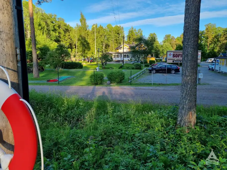 Bild av Sofielund vandrarhem & camping, med en grönskande trädgård omgiven av stora träd och gräsmatta. Skuggiga områden under träden skapar en lugn och inbjudande miljö, perfekt för rekreation och avkoppling. En uppfart leder in mot området, där park- och trädgårdslandskapet ger känsla av en välskött bakgård.