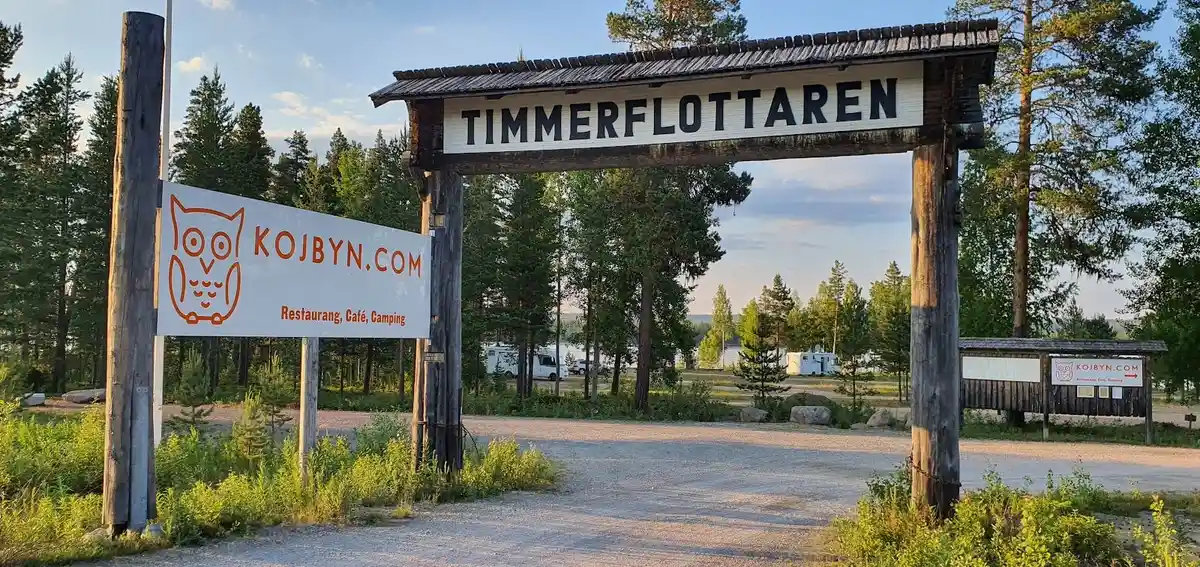 En träskylt märkt "Kojbyns Camping" omgiven av höga barrträd som granar och tallar i en naturskön skogsmiljö.