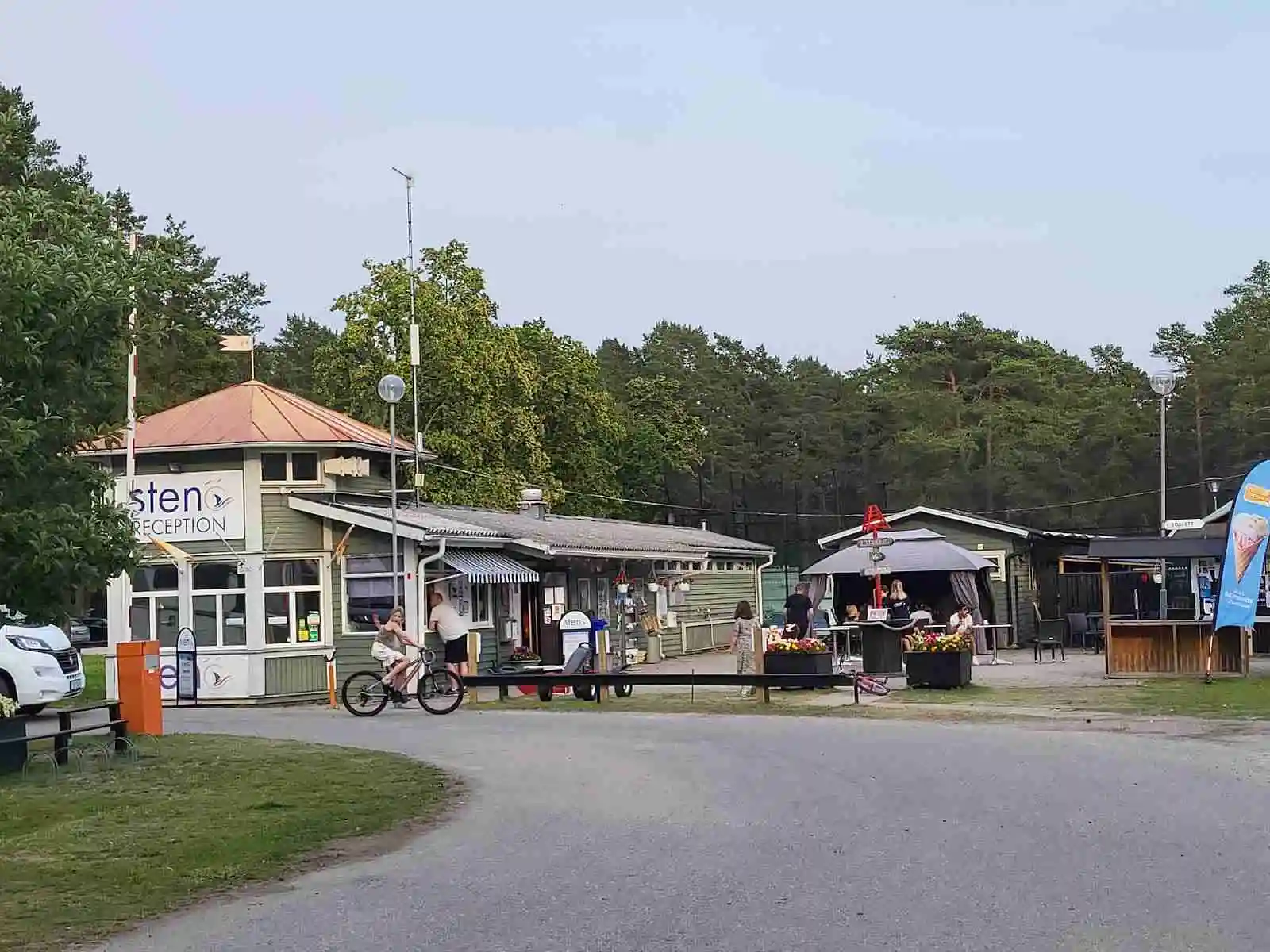 En solig dag på Stenö havsbad & camping, med ett öppet rekreationsområde där människor njuter av fritidsaktiviteter. Ett område för vila med sittplatser och stolpar som markerar olika zoner, omgiven av grönska och en avkopplande atmosfär.