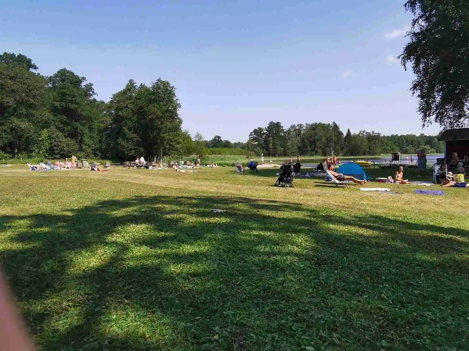 En solig sommareftermiddag på Österby camping med människor som solar, njuter av en picknick och kopplar av i det gröna gräset nära en glittrande sjö.