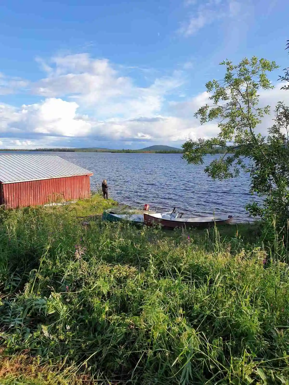 En pittoresk camping vid Piilijärvi med små stugor och skjul vid sjöns strand, omgivna av moln och naturskön landskap.