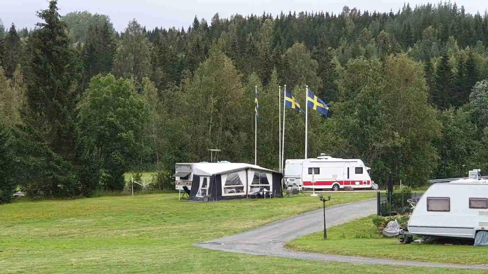 Bild av Docksta Vandrarhem & Camping Kustladan med grönskande ängar och gräsmattor. Flera husvagnar och en husbil står parkerade runt området, omgiven av frodig natur och en parkmiljö som bjuder in till camping och rekreation.
