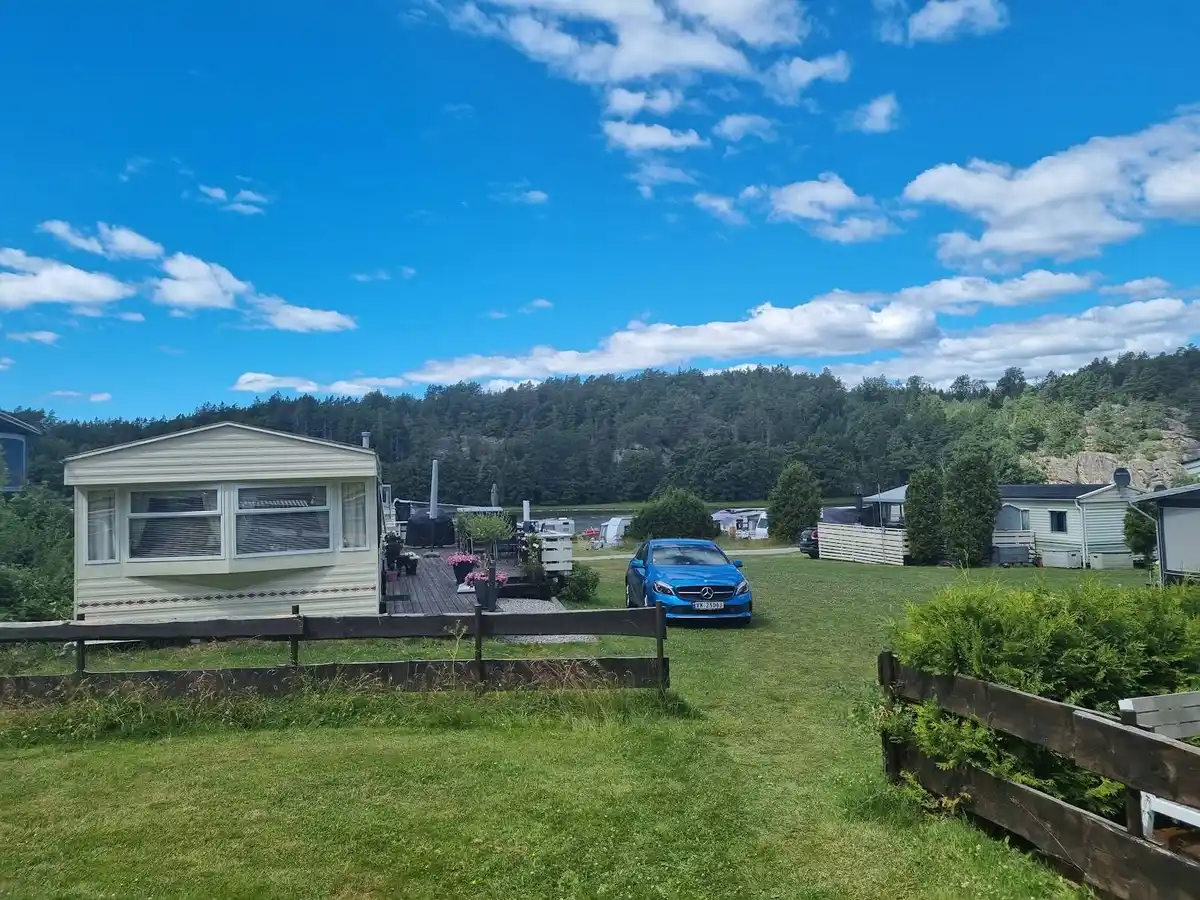 En vidsträckt campingplats med flera husvagnar parkerade på den gröna gräsmattan. I bakgrunden syns ett hus och en klar blå himmel med vita moln.