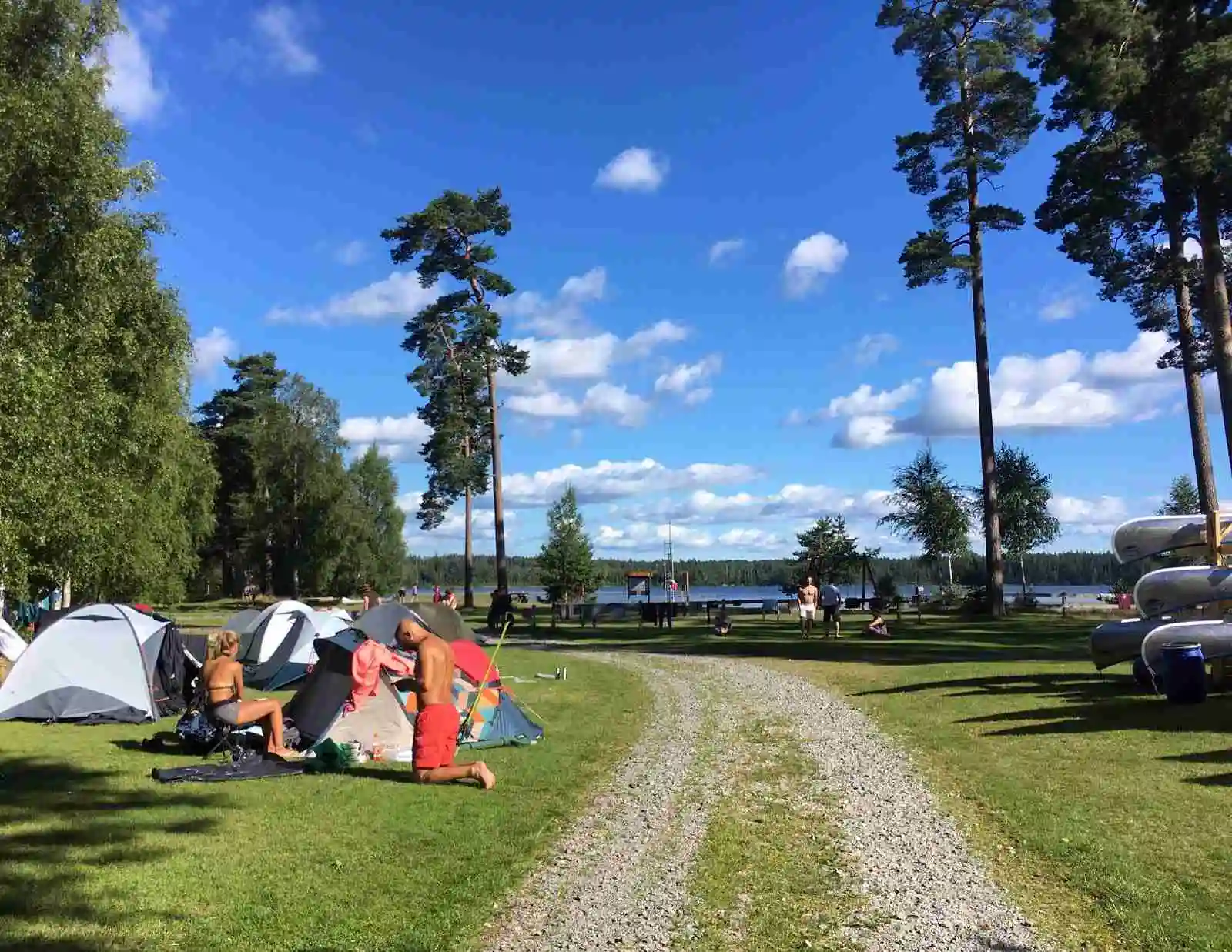 En campingplats vid en sjö med tält uppsatta på en gräsyta, omgiven av frodig grönska och under en himmel med cumulusmoln.