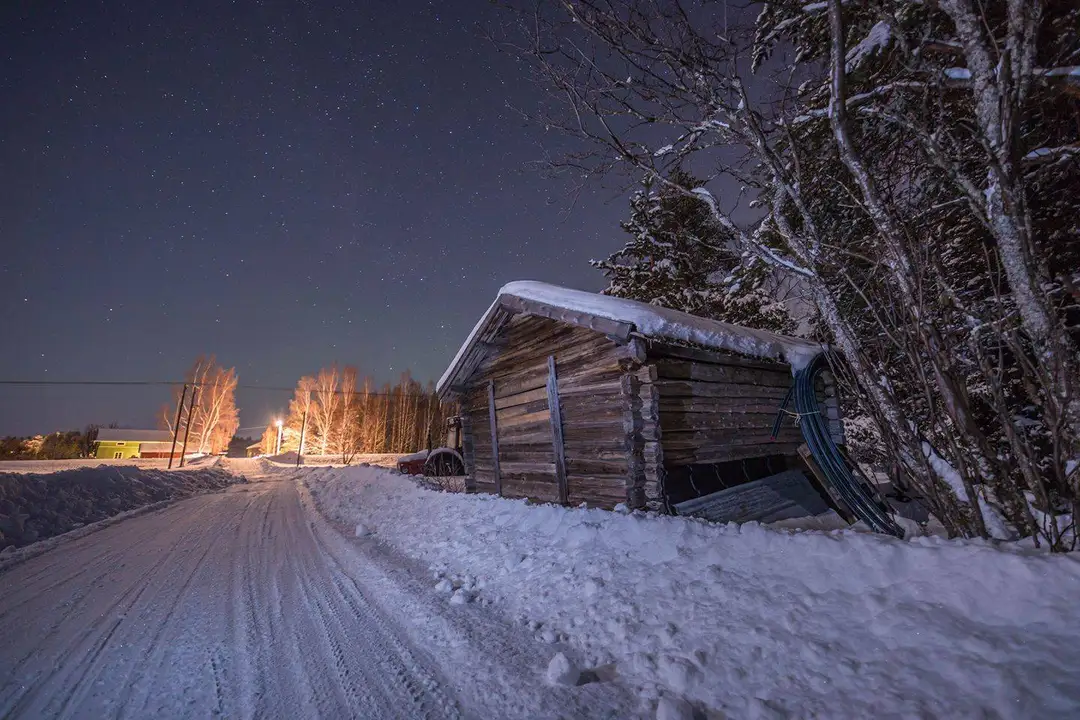 En vinterbild av ett snötäckt landskap vid Seskarö Havsbad & Camping, med en liten stuga omgiven av snötyngda grenar under natthimlen.