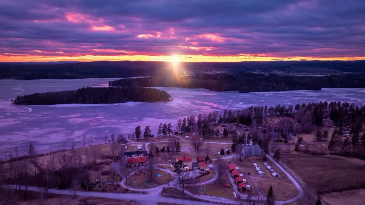 En vintrig campingplats under skymningen, täckt av snö med en färgsprakande solnedgång i bakgrunden.