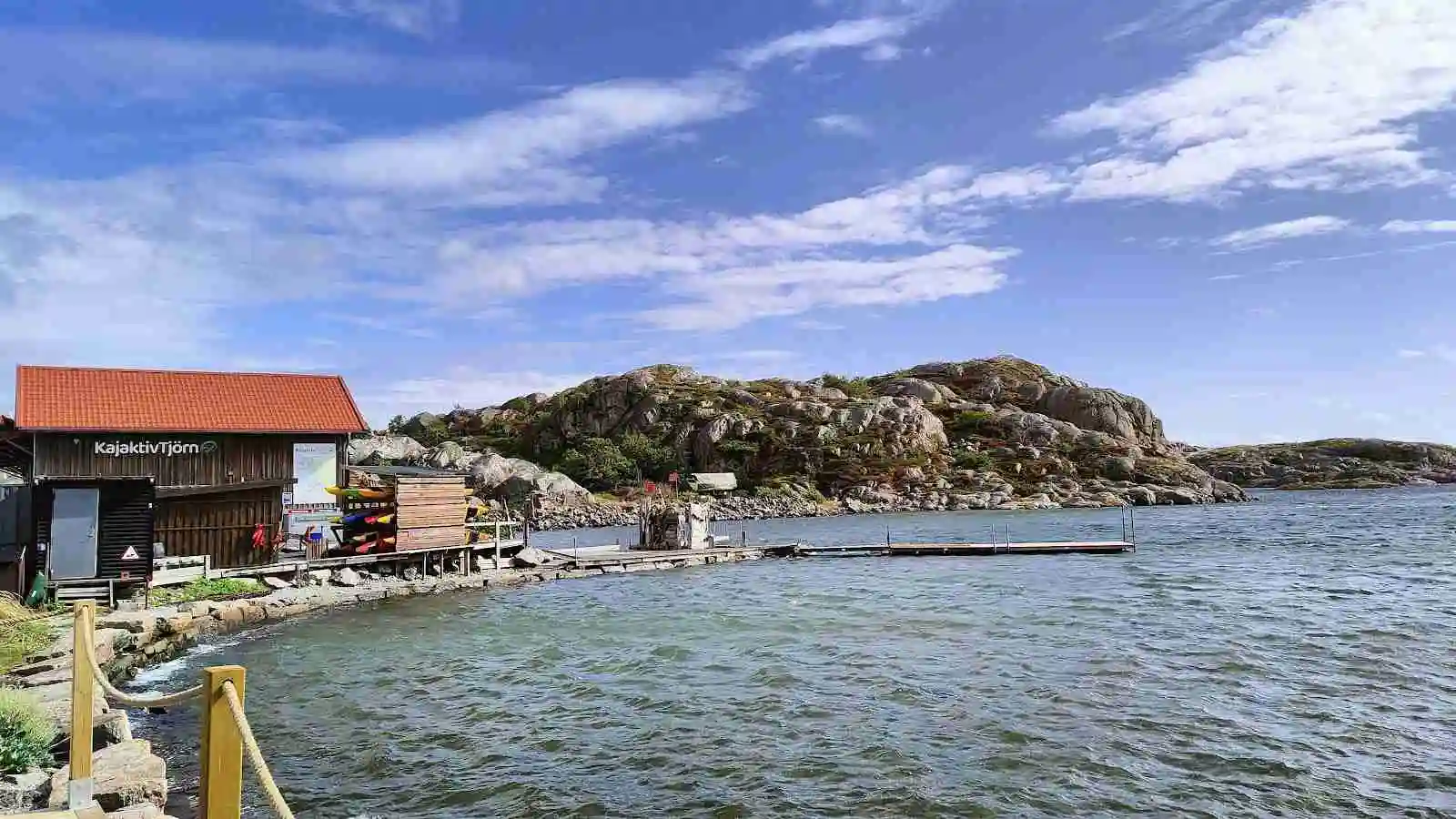 Bild på Mossholmen med vatten i förgrunden och en klippig kustlinje. I bakgrunden syns små stugor, en ö och havet.