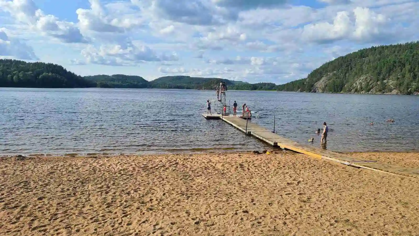 En lugn sommardag vid Pinnarp Camping, med en bred sjö som glittrar i solskenet. Sandstranden löper längs kanten av vattnet, omgiven av grönska och inbjuder till bad och avkoppling.