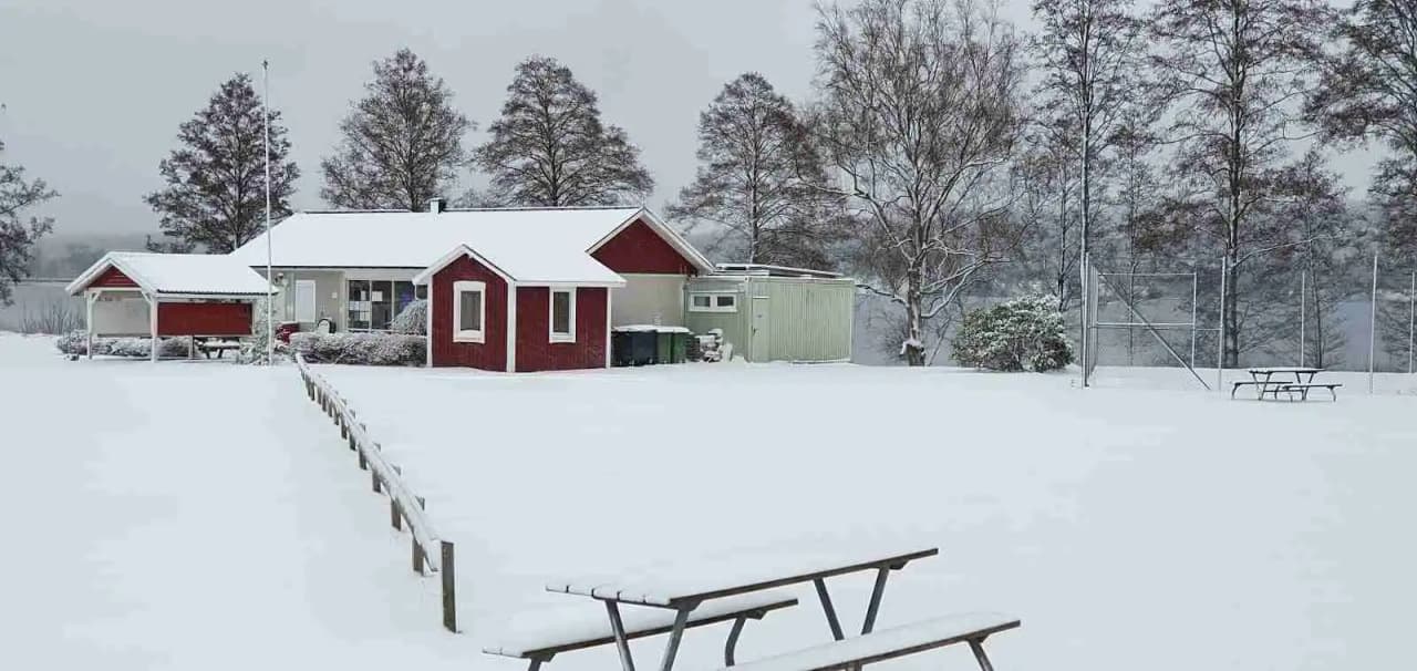 En vinterbild av Ramkvilla Camping med snötäckta stugor omgivna av snötäckta trädgrenar. Stugornas tak är täckta av ett tjockt lager snö, vilket bidrar till en mysig och fridfull atmosfär i detta vinterlandskap.