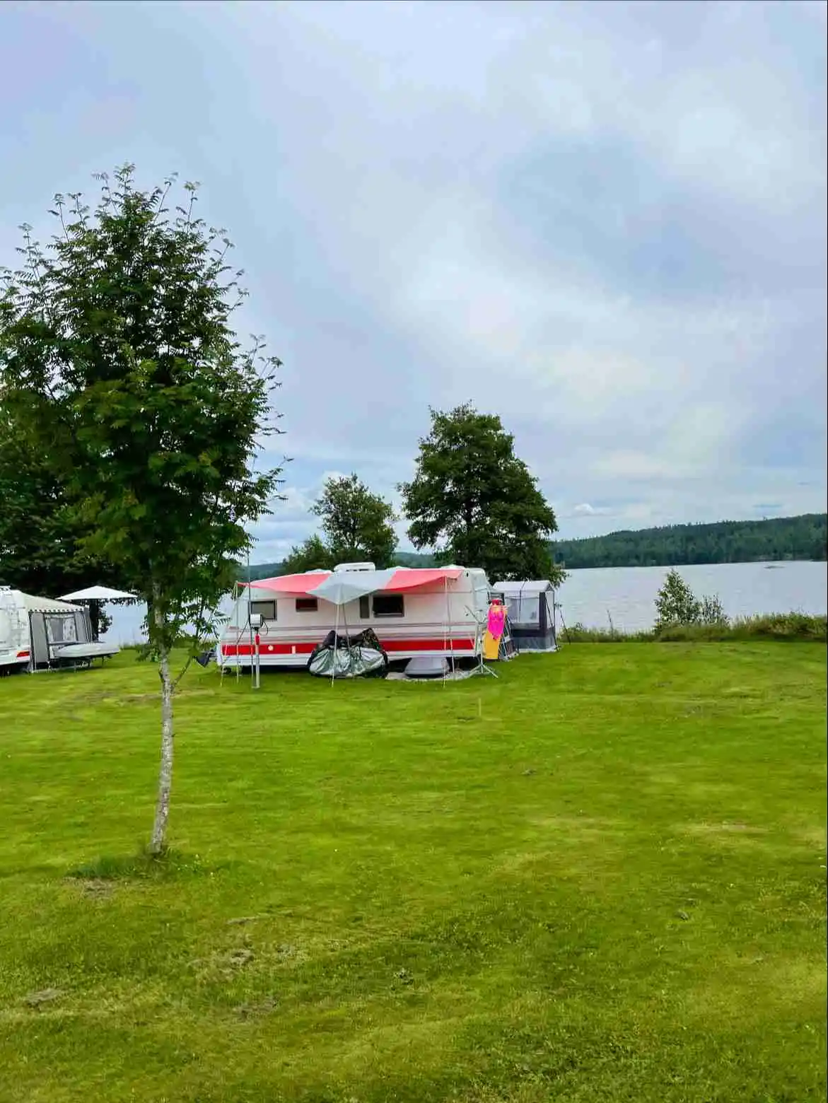 En grön campingplats i Ramkvilla med husbilar parkerade på en välskött gräsmatta. Moln syns på himlen och en närliggande sjö kan skymtas i bakgrunden, omgiven av en lummig park.