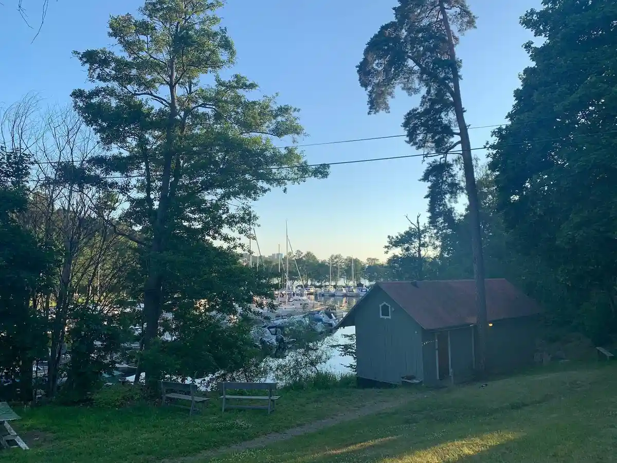 En båt med mast ligger förtöjd vid en grönskande park, skuggad av stolpar och träd. I bakgrunden syns en vattenväg som ger ett lugnande intryck.