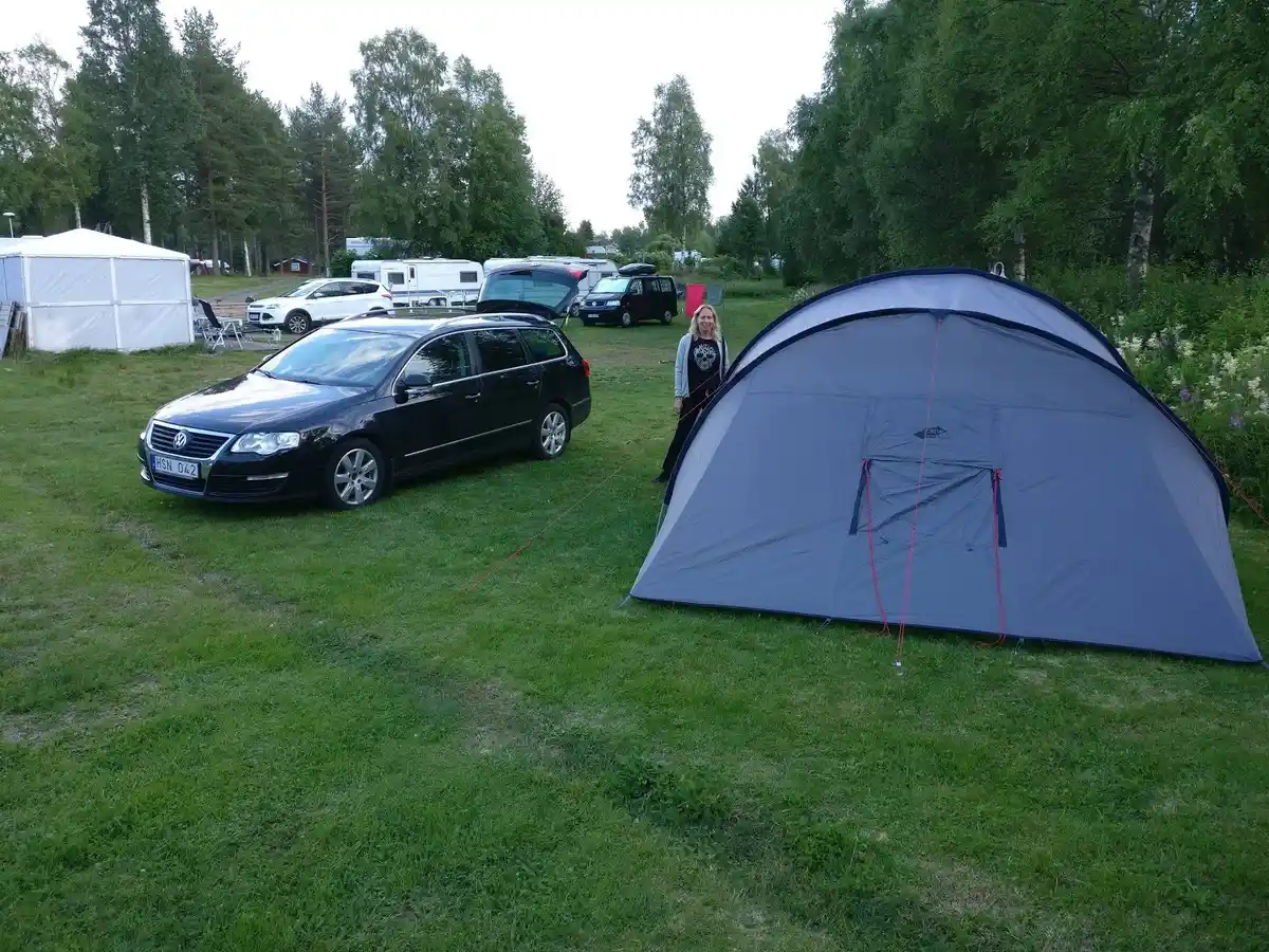 En campingplats med tält och en bil parkerad i närheten, omgiven av skog och natur. Packning och vandringsutrustning syns bredvid tältet.