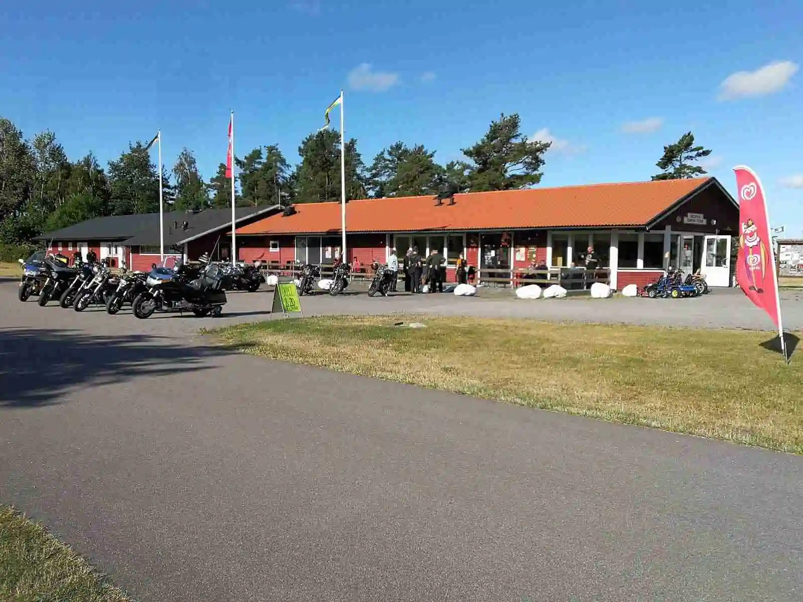 En campingplats med en motorcykel parkerad vid en stolpe på en asfalterad yta, omgiven av grönska. 