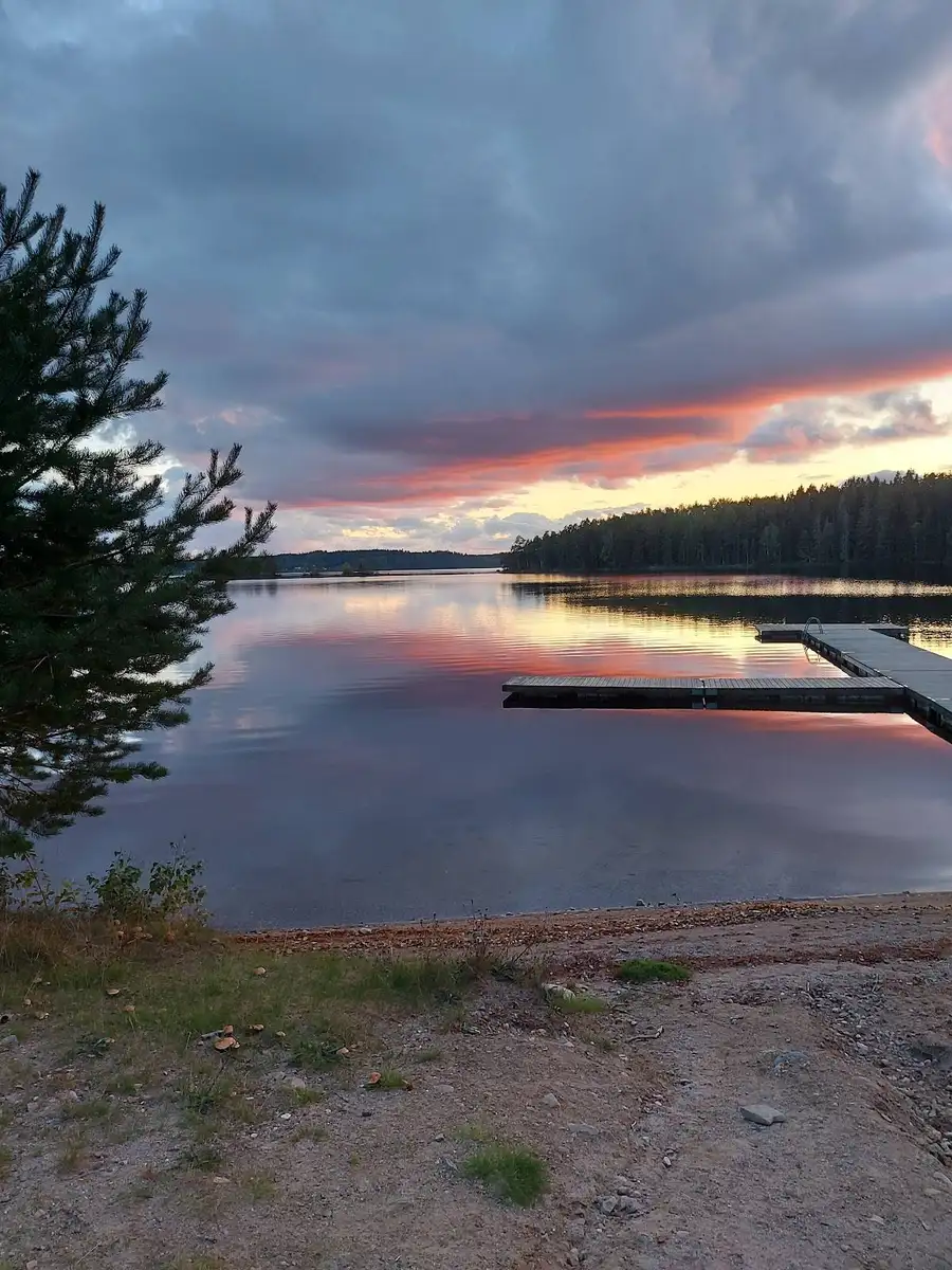 En sjö vid solnedgången med lugnt vatten och ett färgglatt sken på himlen, omgiven av träd och naturlandskap på Uskavigårdens camping.