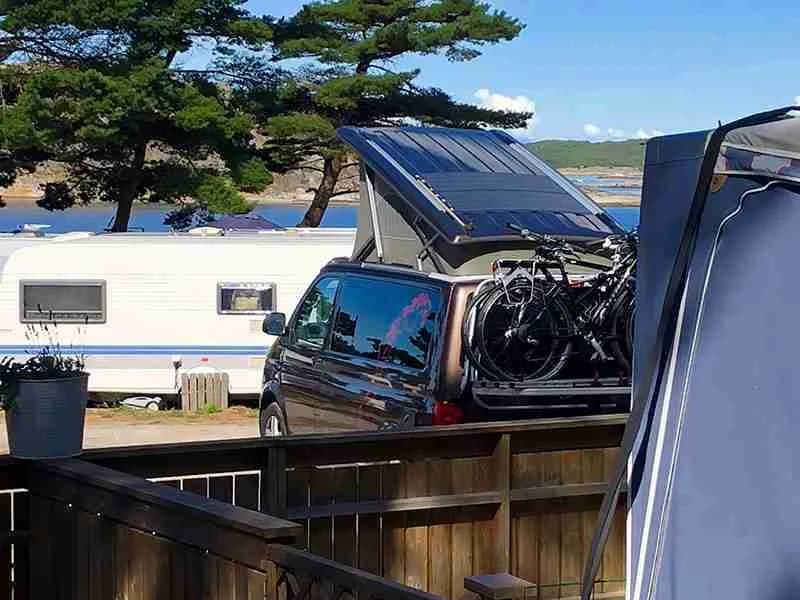 En campingplats med flera parkerade bilar och husbilar, tydliga bilars bärgningssystem och baklyktor syns. I bakgrunden finns ett staket.