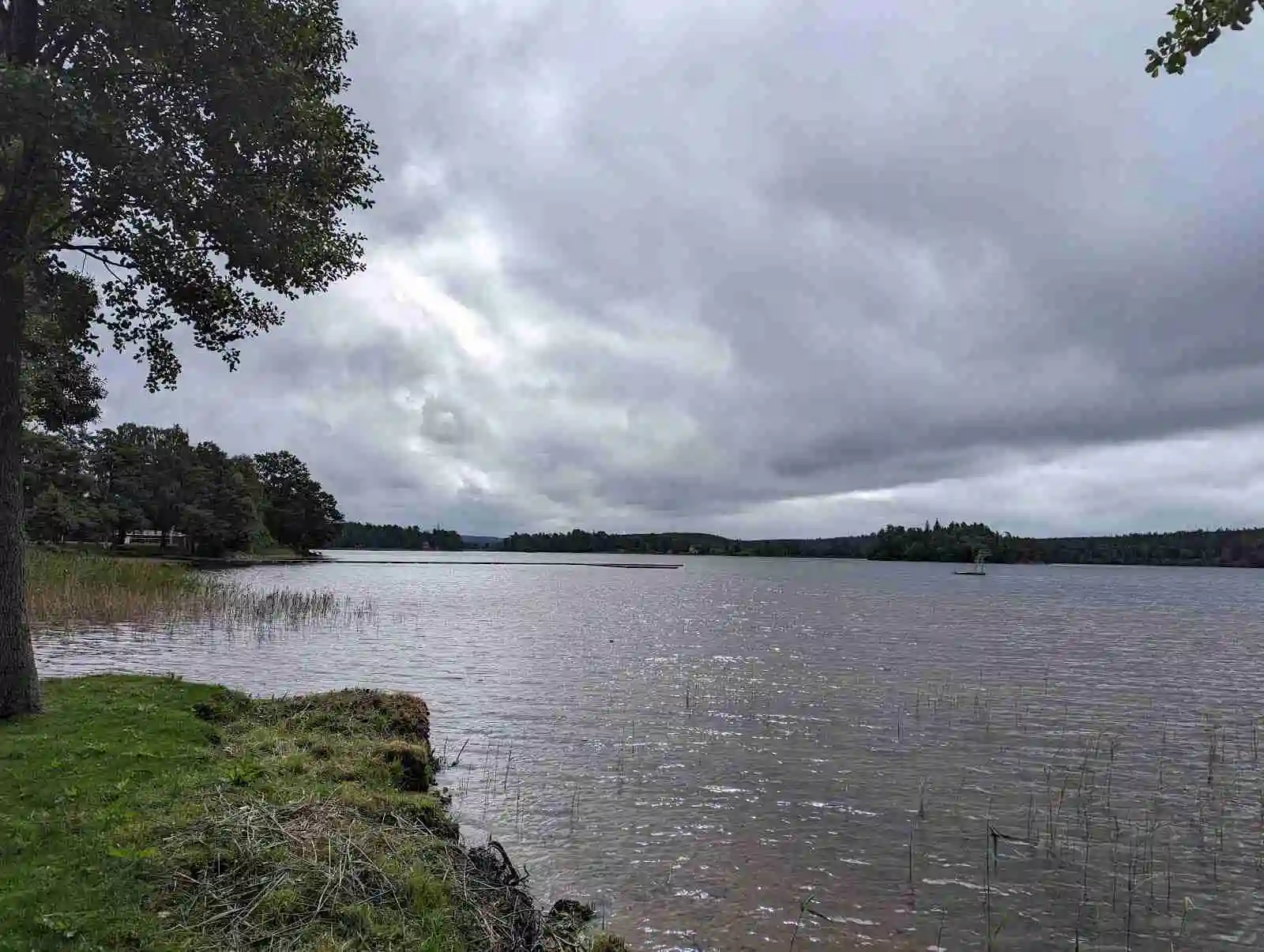 En naturskön vy över Ramkvilla camping med en lugn vattenspegel i förgrunden och en klar himmel med moln i bakgrunden.