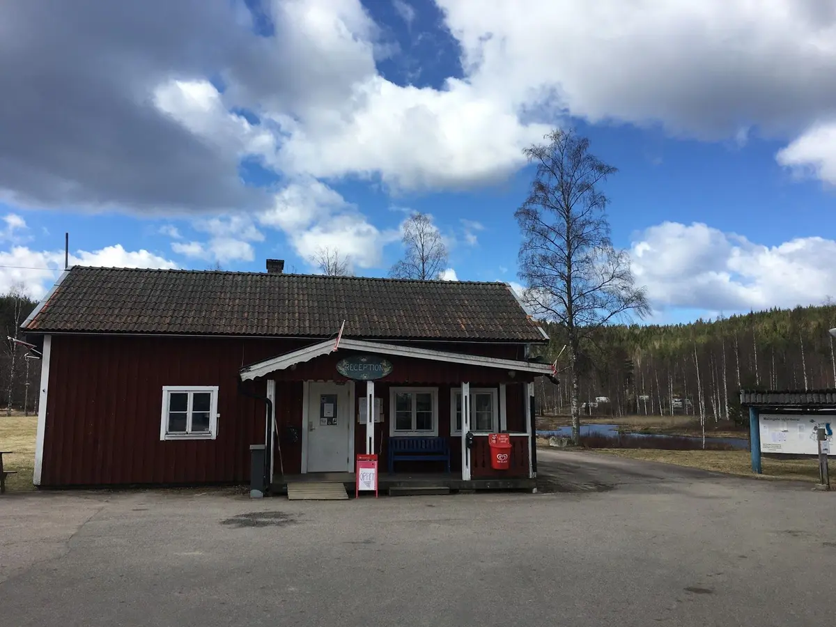En asfalterad infart med en skyltstolpe och en väg som leder mot några enkla byggnader på Malingsbo camping, omgivna av grönska.