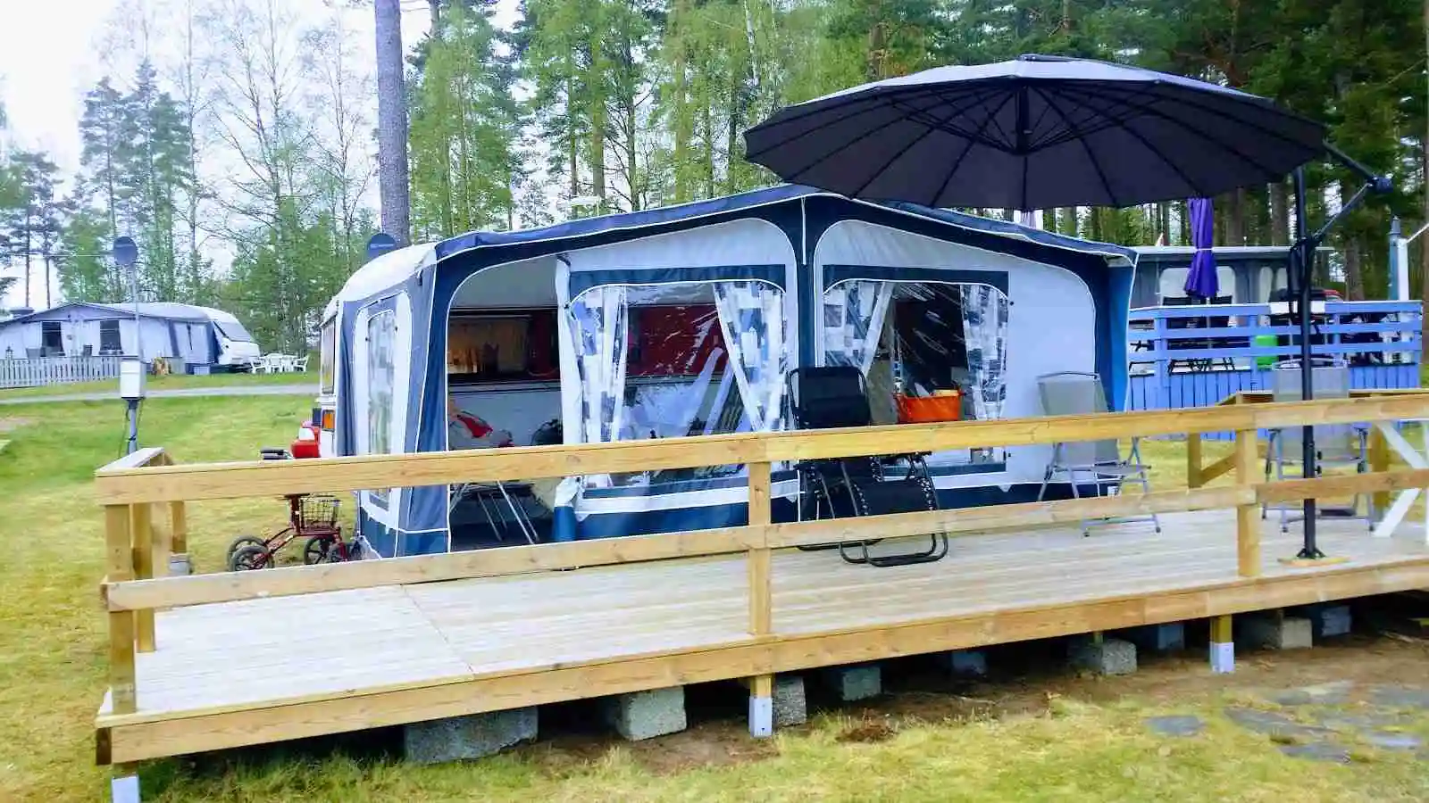 Bild av en campingplats på Treens natur & fiskecamp, med en husvagn uppställd i skuggan. Framför husvagnen finns utemöbler på en träveranda, omgiven av frodig grönska.