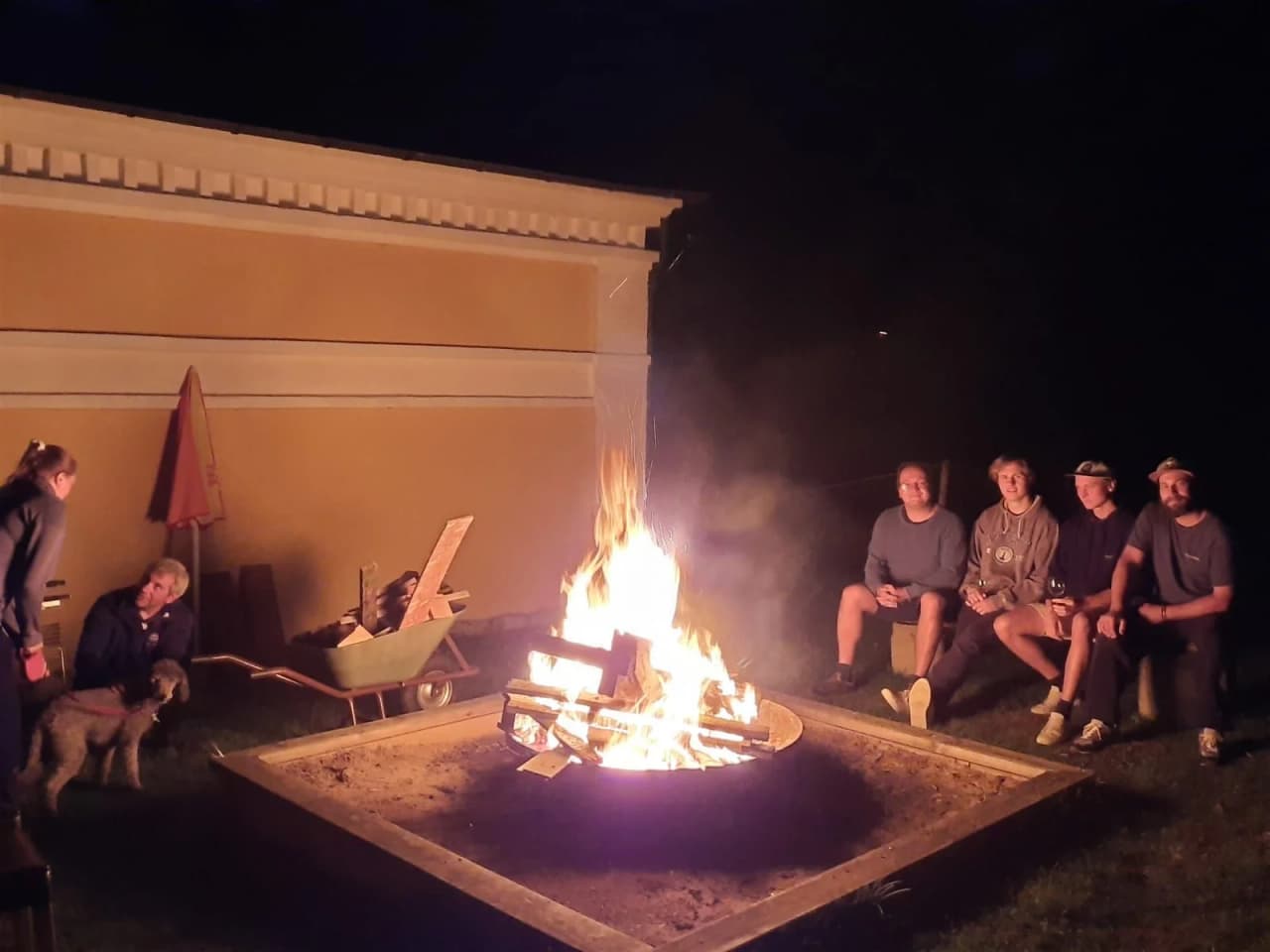 En grupp människor sitter runt en lägereld vid Hunnebergs Gård Hostel & Camping, omgivna av campingstolar, i en avslappnad och trevlig atmosfär.