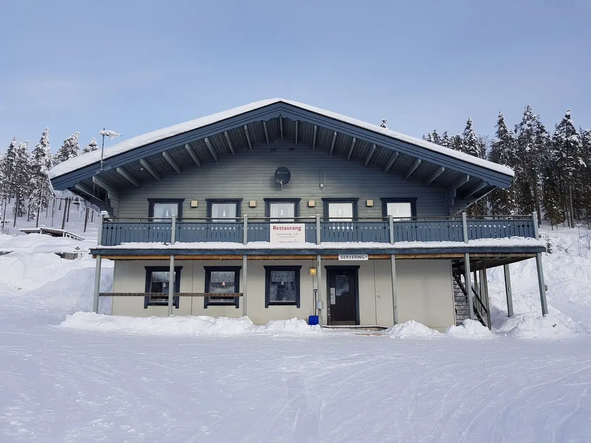 Ett vinterlandskap vid Storklinta Skidcenter AB med ett snötäckt hus i förgrunden, omgiven av snöiga backar och vinterklädda träd.