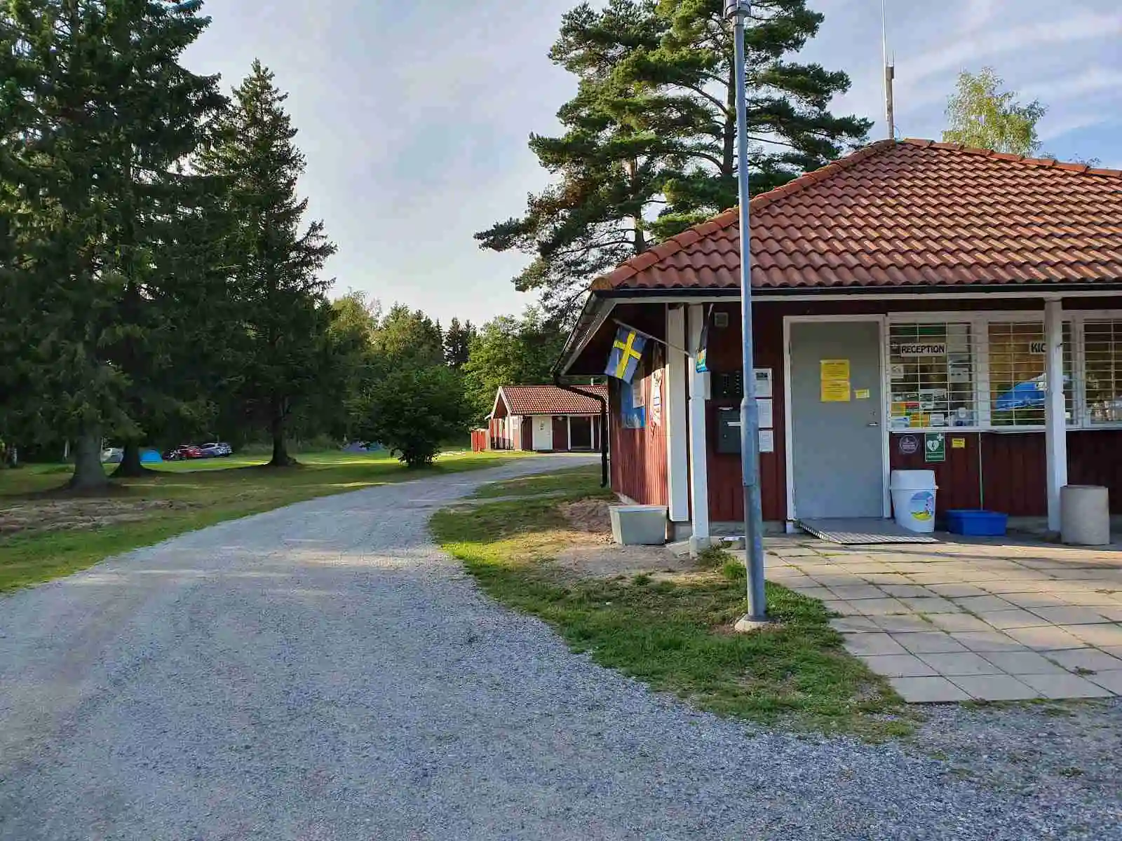 En grönskande campingplats med flera små stugor utspridda runt området. Stugorna har bruna tak och ligger på en välskött gräsmatta. Ett par skuggföreställningar syns från träd eller stolpar i närheten. En gångväg löper genom området och det finns öppna ytor där besökare kan umgås.