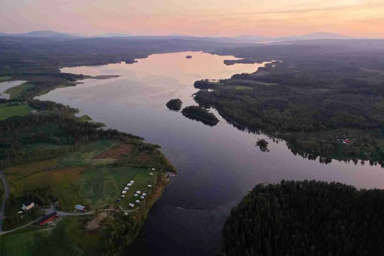 Bild av Kvissleströmmens fiskecamping: En naturskön vy över en bred flod eller sjö med lugnt vatten, omgiven av grönskande träd och vegetationsrika stränder. Stämningen är fridfull och inbjuder till fiske och avkoppling i en vacker naturlig miljö.