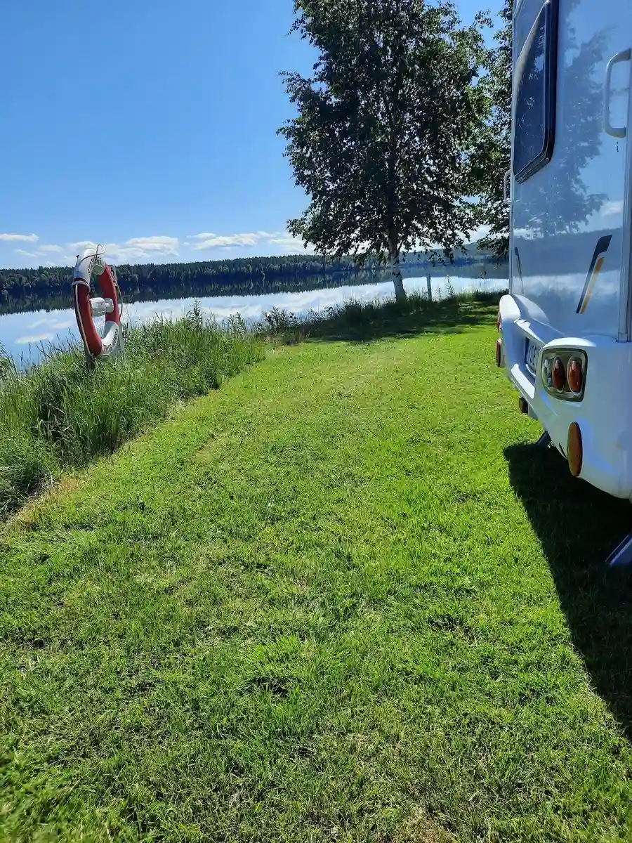 En husvagn står parkerad på en gräsbevuxen campingplats vid Vindelforsarna, omgiven av gröna ytor och träd.