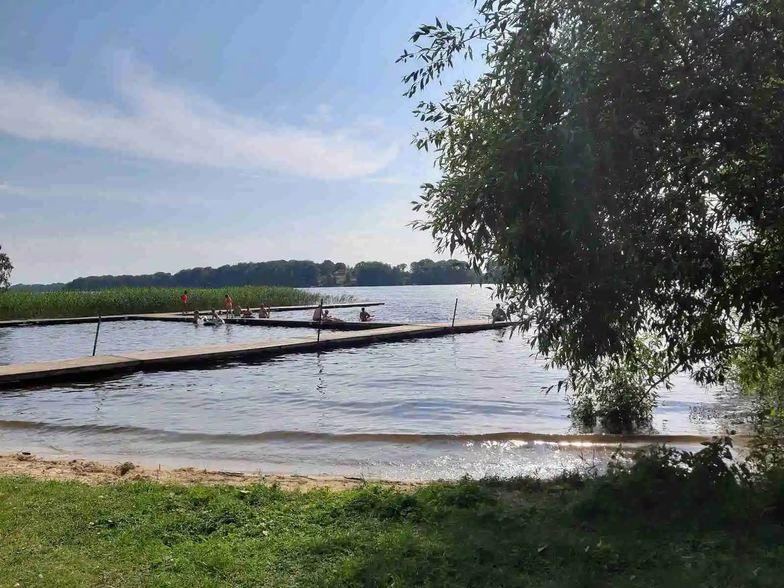 En somrig bild av Ivö Camping vid en sjö med en brygga, omgiven av lummig natur och en stilla vattenyta, idealisk för rekreation och friluftsliv.