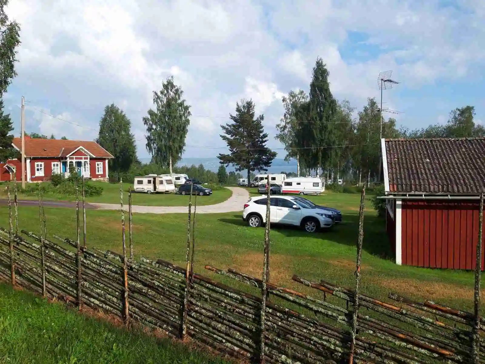 Bild av Vikarbygården: En pittoresk landsbygdsscen med en stuga omgiven av grön gräsmatta och växter. Moln syns på himlen över ett öppet landskap, vilket skapar en rofylld och lantlig atmosfär.