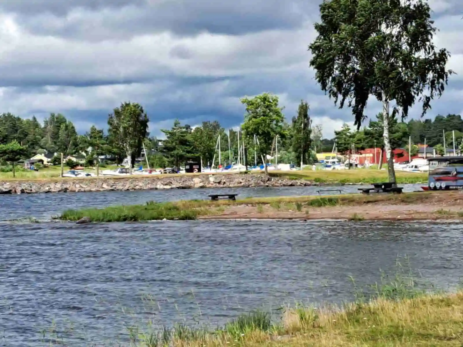 En somrig bild av Malexander Camping visar en stilla vattendrag med grönskande strandkanter. Moln pryder himlen i bakgrunden, och vattnet speglar det klara vädret.