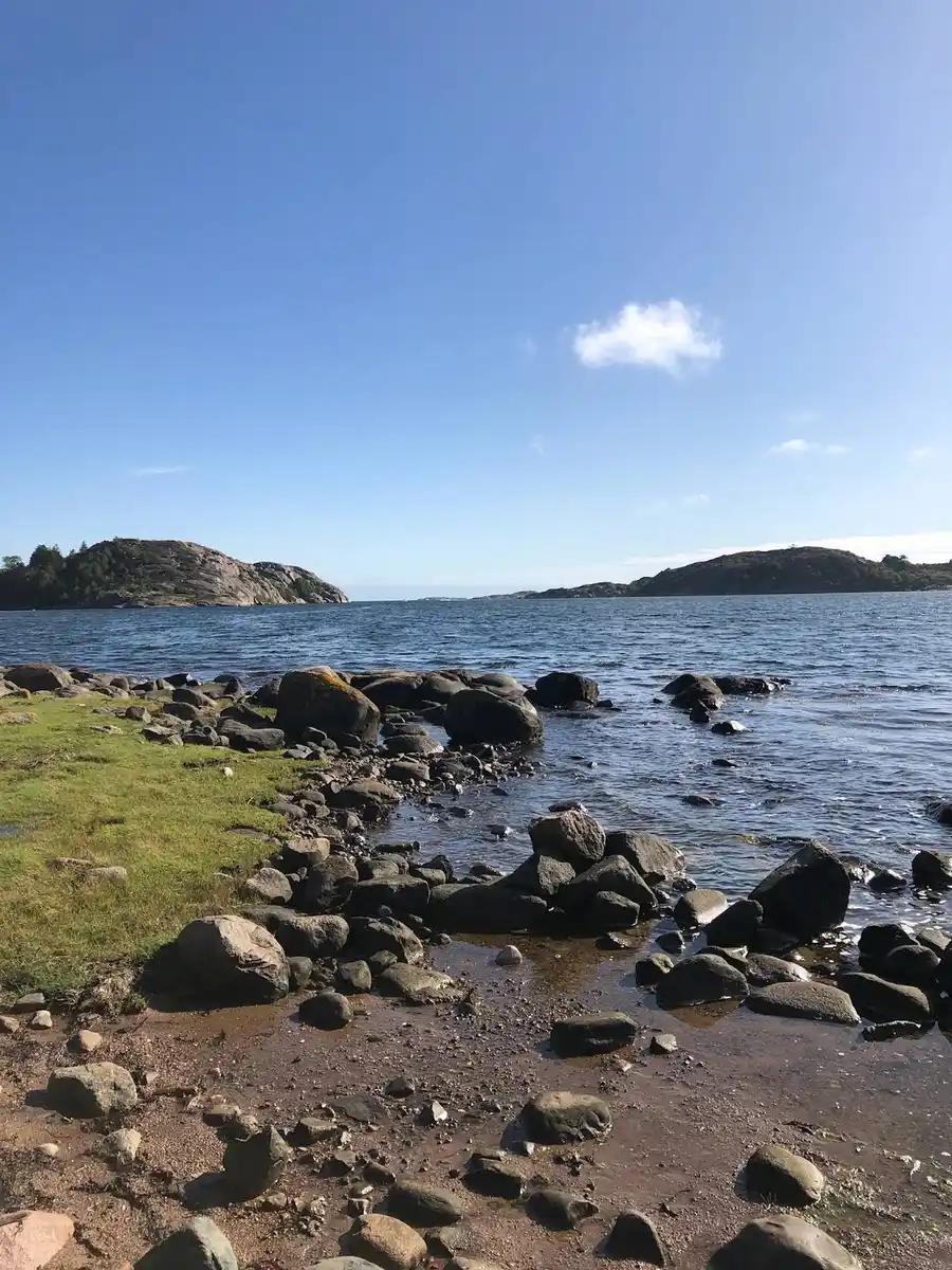 En klarblå himmel sträcker sig över en rofylld kustlinje vid Örns camping, där vatten möter kanten av stenar och klippor. Vid horisonten smälter himlen och vattnet samman, ackompanjerat av några spridda moln.