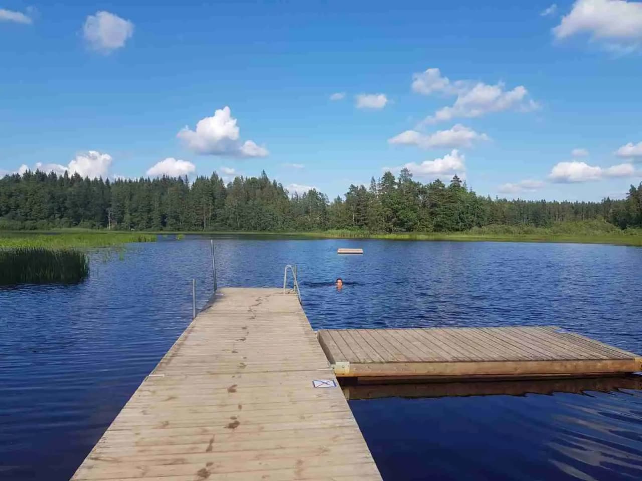 Bild av Emmaboda camping visar en fridfull sjö omgiven av grönska. En träbrygga sträcker sig ut mot vattnet, där man kan njuta av det lugna landskapet.