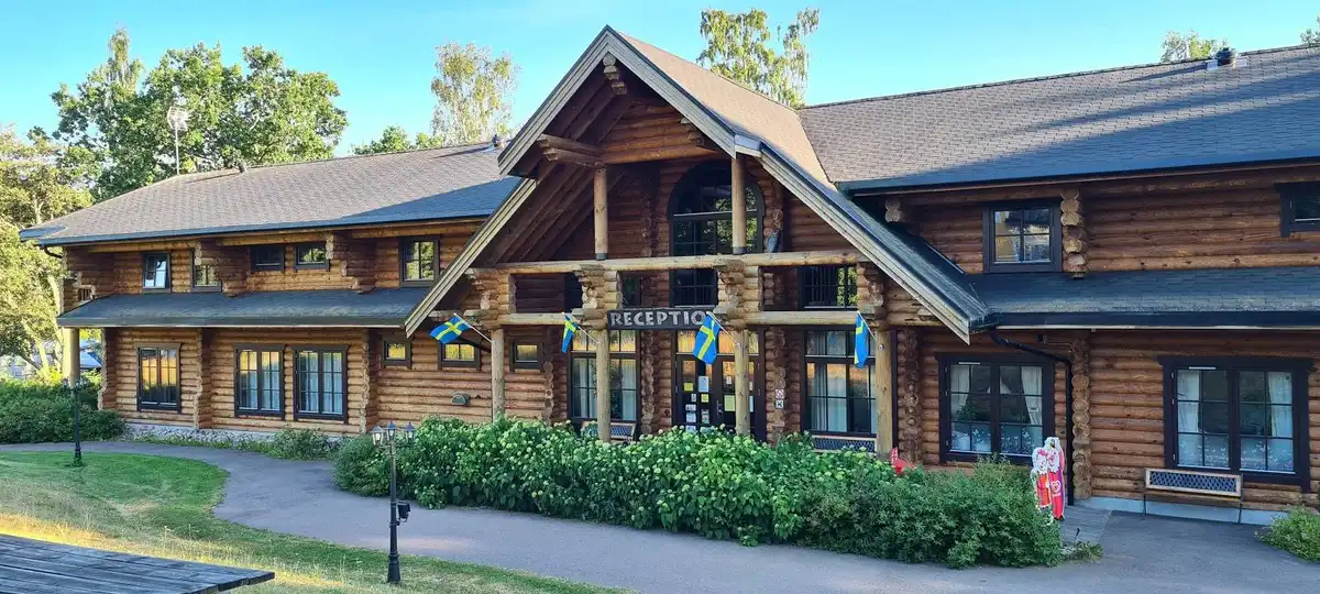 En trästuga med sluttande tak och träpanel, omgiven av grönska och en liten gårdsplan.