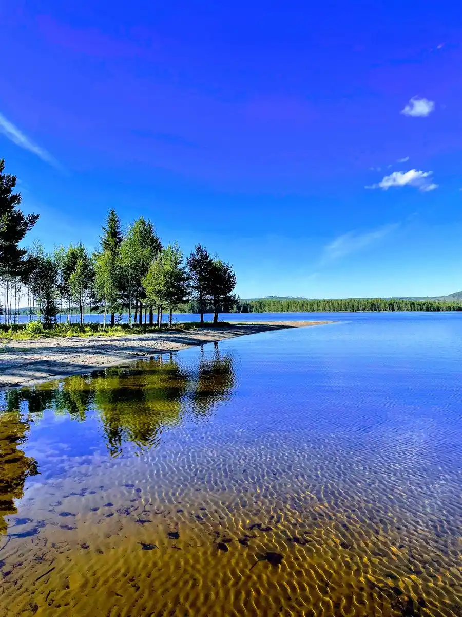 En lugn sommardag vid Kojbyns camping med klarblått vatten, en vidsträckt horisont och moln på himlen som speglas i sjön.