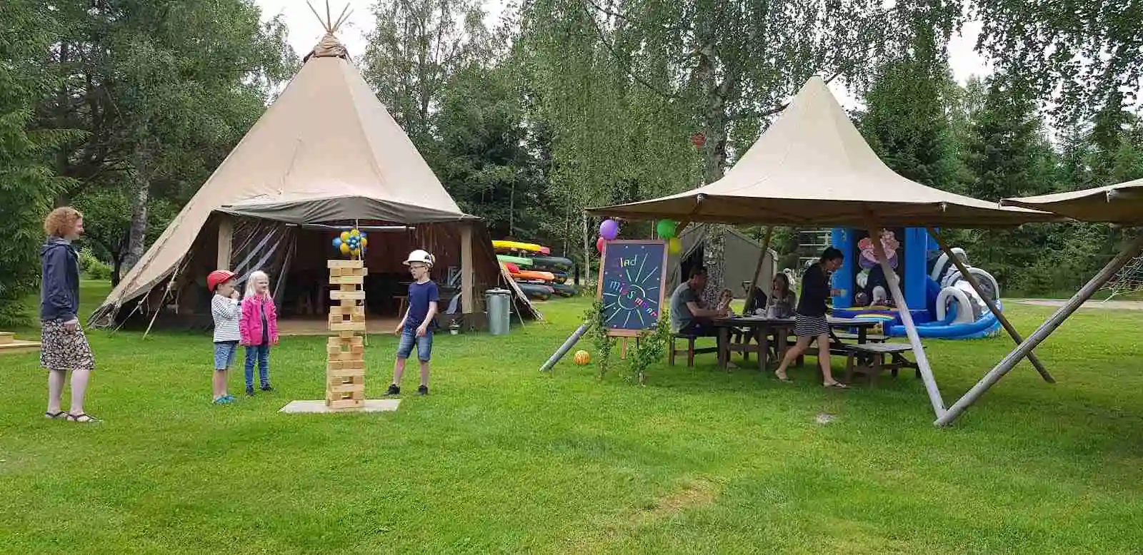 En campingplats i Tiveden med tältuppställning under skugga. Tält och presenningar är uppsatta med stänger, omgivna av grönska och skog. Människor njuter av fritid och rekreation i naturen.