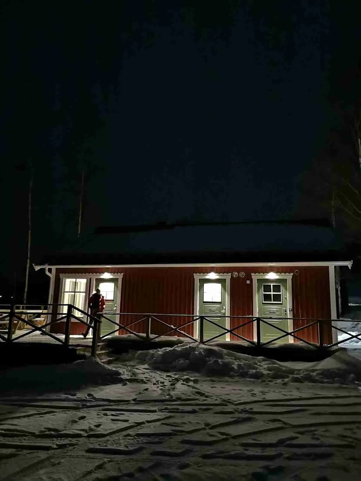 En snötäckt stuga vid Gällstasjöns ställplats under en vinterkväll, upplyst i mörkret.