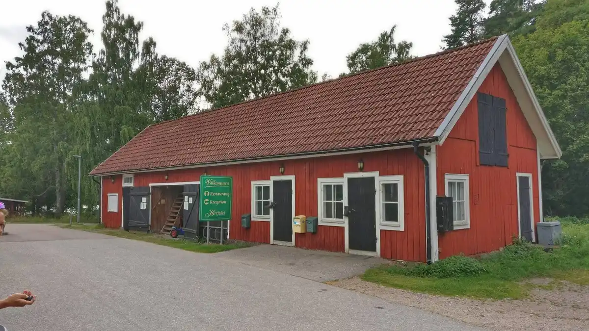 En röd stuga ligger på en grönskande gård med en grusad infart, omgiven av andra mindre byggnader såsom ett skjul och en ladugård.