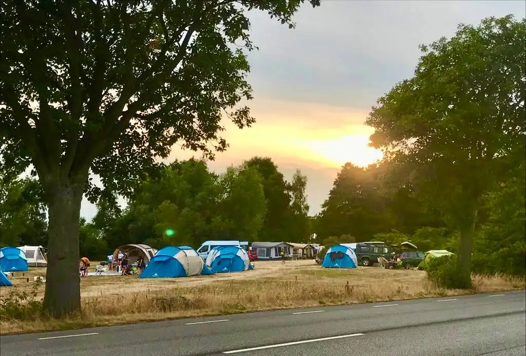 En somrig campingplats vid Lomma camping med tält uppsatta på gräset, och ett par bilar parkerade i bakgrunden under en himmel med cumulusmoln. En vit skåpbil står vid sidan om tältområdet.