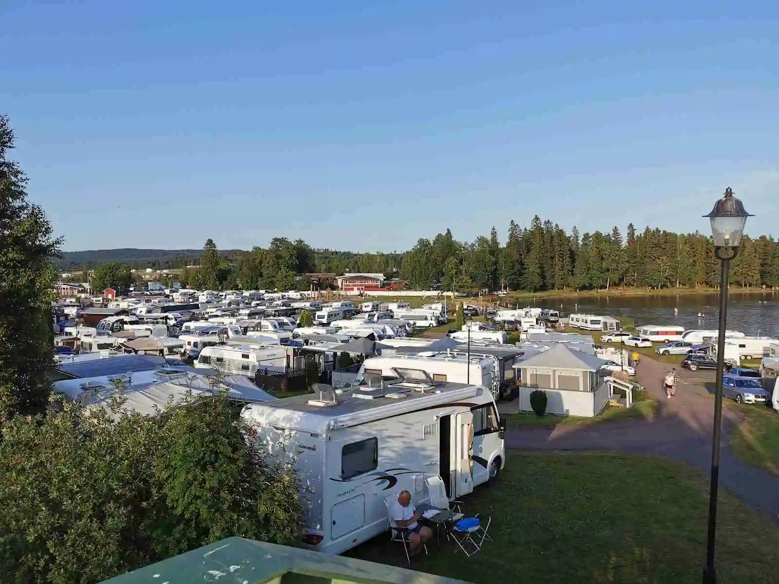 Flygbild över Sunne Camping och Sommarland, visar en stor parkeringsplats med flera husbilar och husvagnar, samt omgivande semesteranläggning i en naturskön miljö.