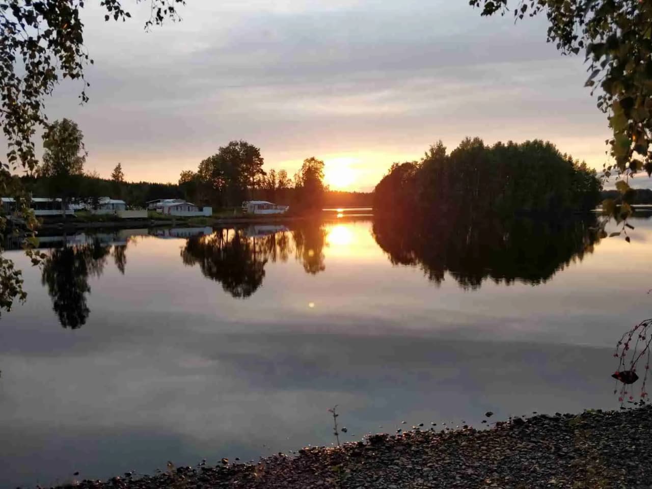 Vy över Krokomvikens camping där en vacker vattenyta reflekterar den stämningsfulla himlen vid skymningen, omgiven av natur och grönska.